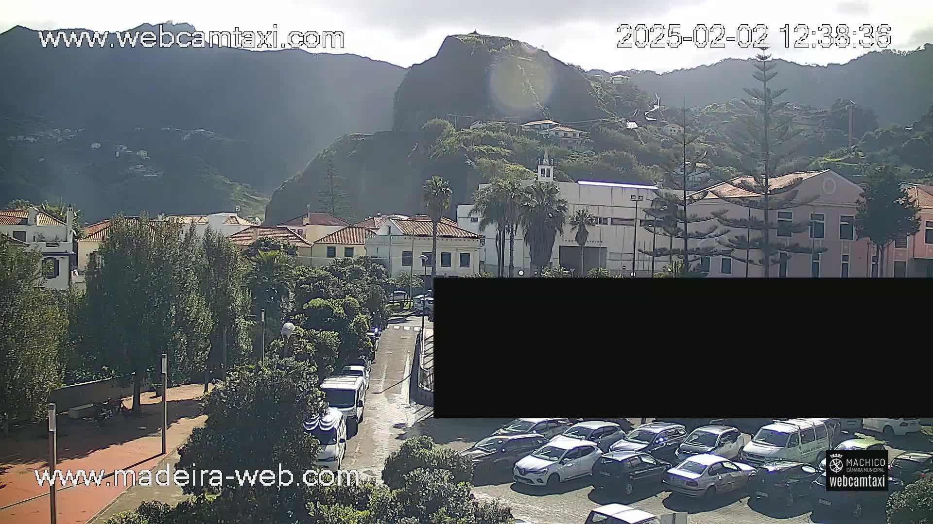 A sunny day reveals a coastal town nestled against mountains, with parked cars lining a street and buildings in the foreground.