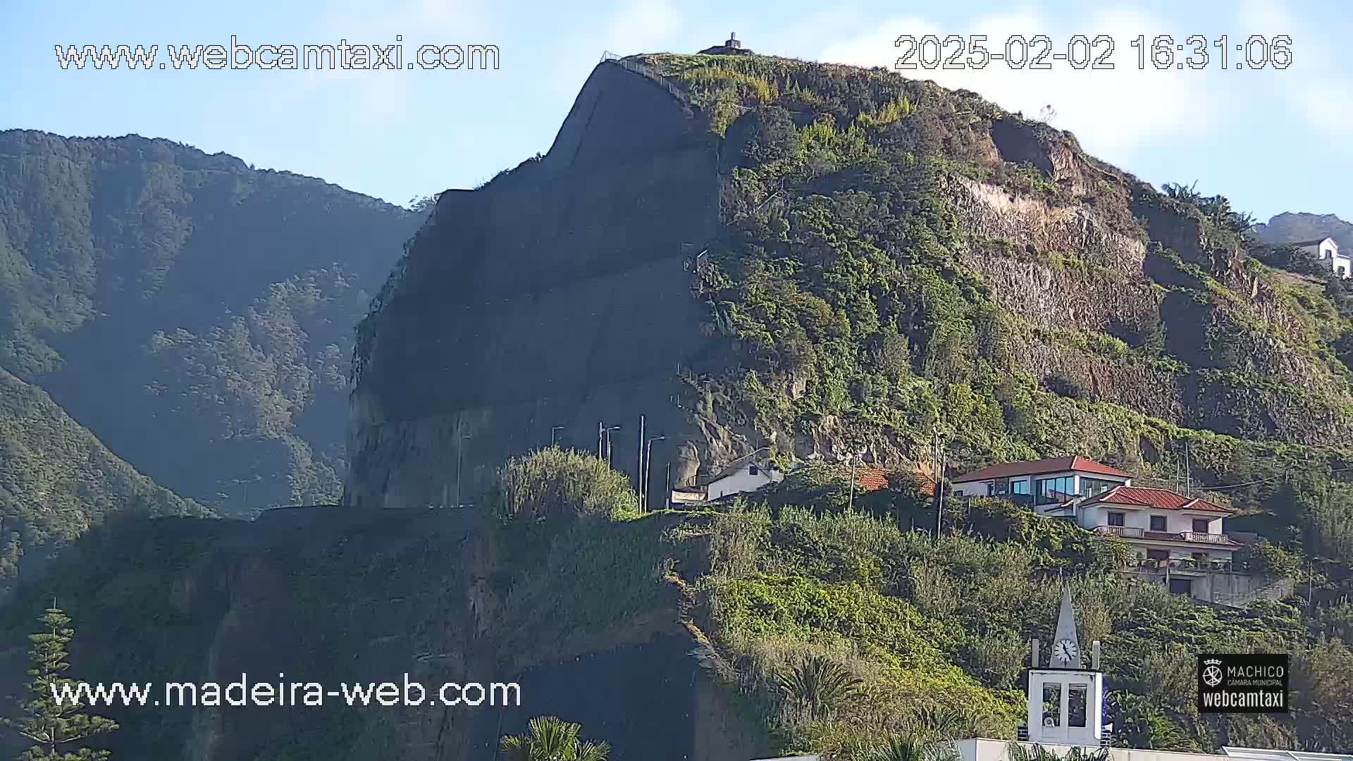 A steep, verdant hillside, partially reinforced with dark material, overlooks a small cluster of houses under a clear sky.