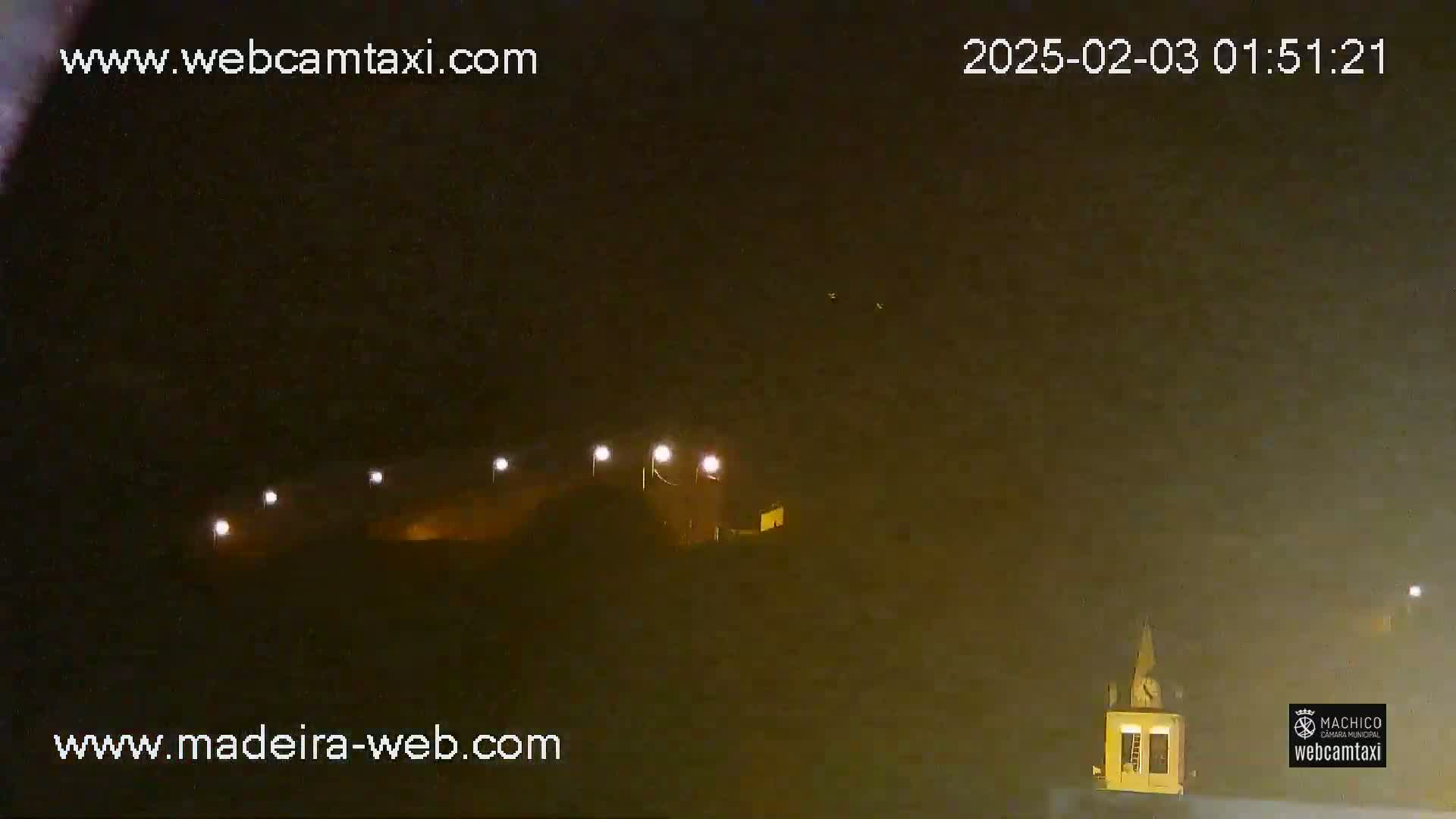 A nighttime, hazy view shows a low building with lights illuminated, and a tall structure with a clock visible in the distance.