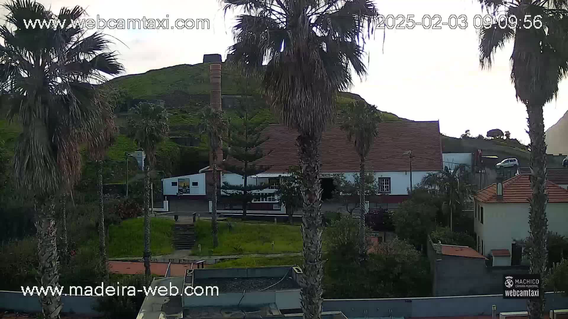A partly cloudy day reveals a hillside village scene with palm trees, low buildings, and a stone structure resembling an old factory or mill.