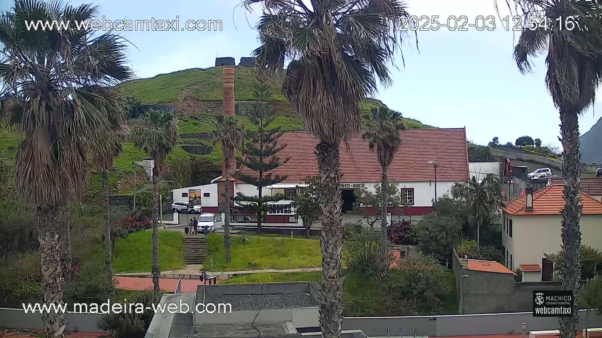 A partly cloudy day reveals a hillside village with palm trees, a white building with a red tile roof, and a brick chimney.