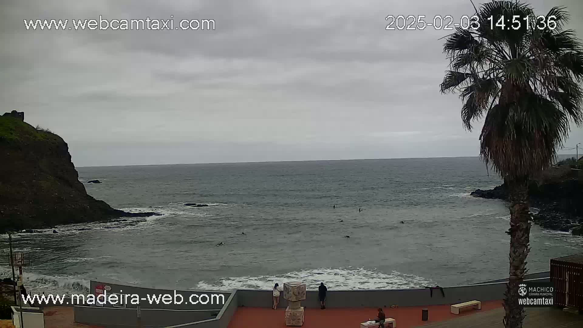 A cloudy day shows a rocky coast with a few people standing near the ocean, where several people are swimming or surfing.