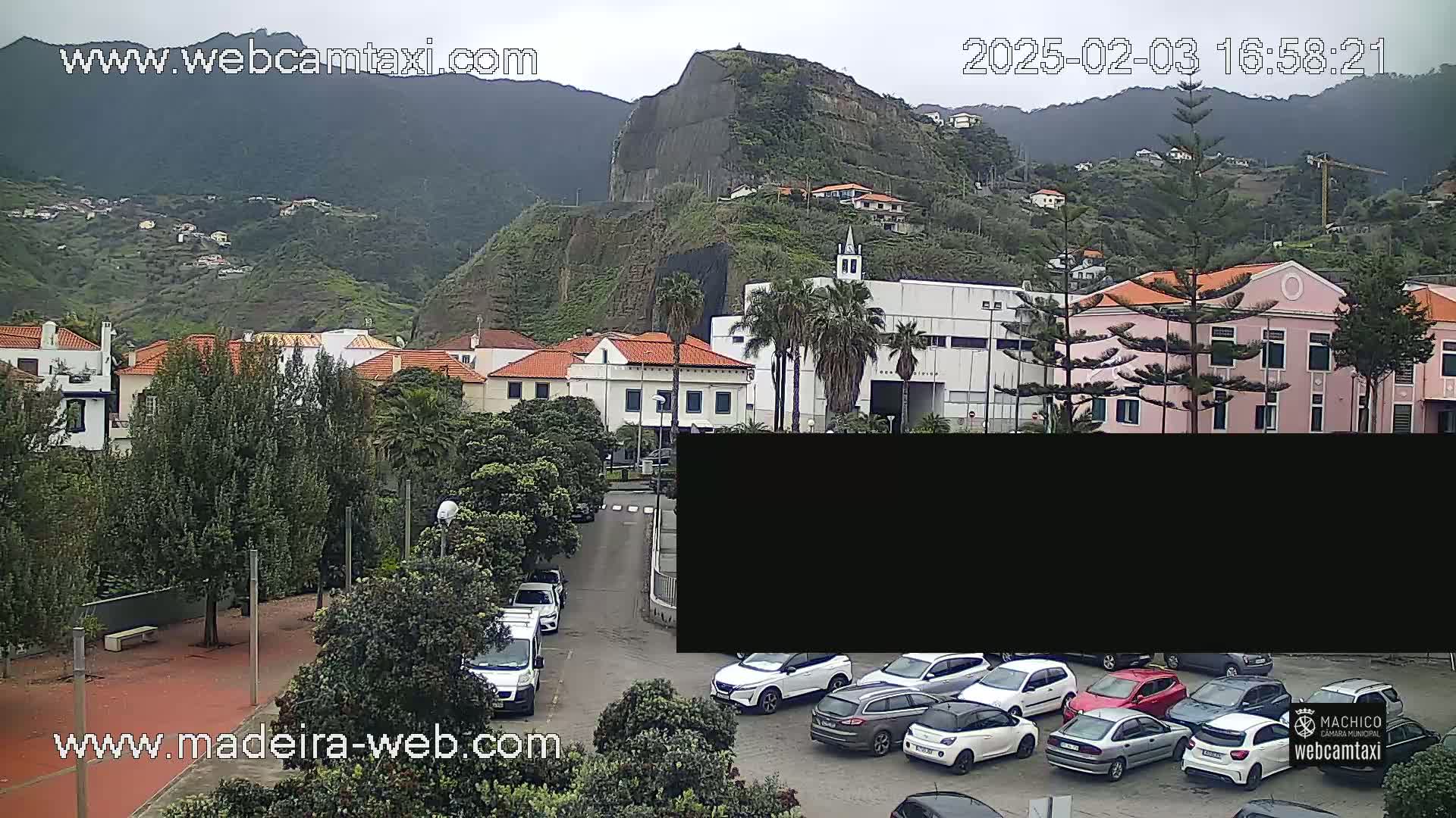 A town nestled against a mountainside under an overcast sky, with several cars parked in a lot and a few driving on a nearby road.