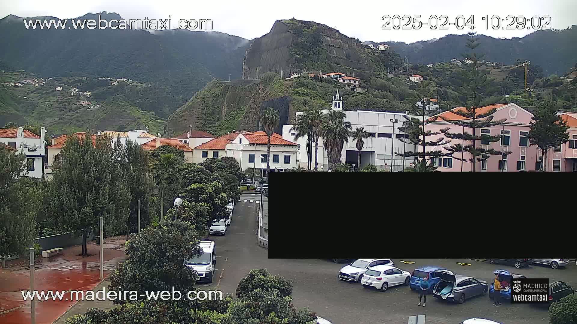 A parking lot with several cars is seen in front of a row of buildings at the base of a large, green, mountainous area under an overcast sky.