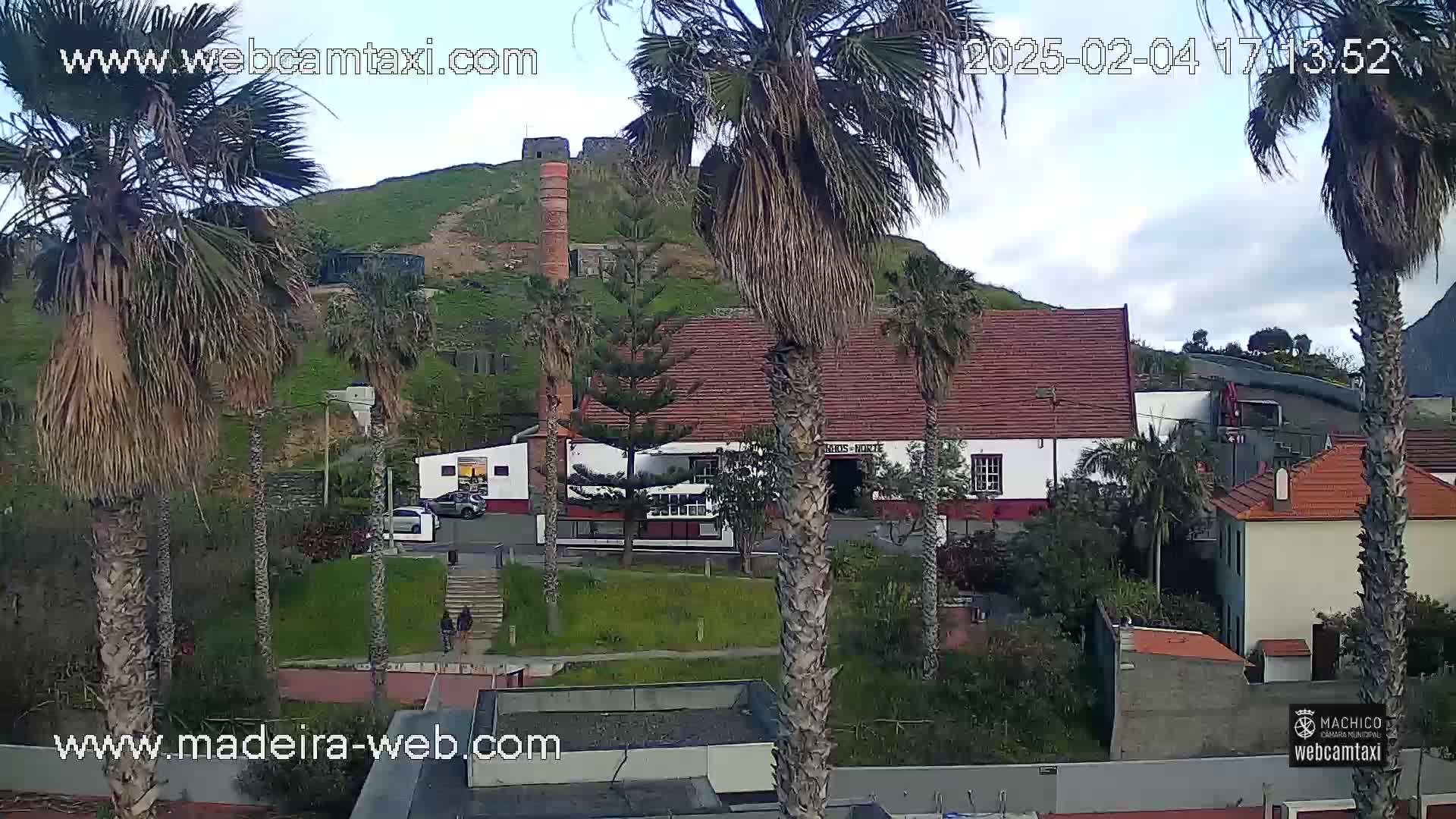 A partly cloudy day reveals a hillside with a red-roofed building, palm trees, and a stone structure, all viewed from a slightly elevated perspective.