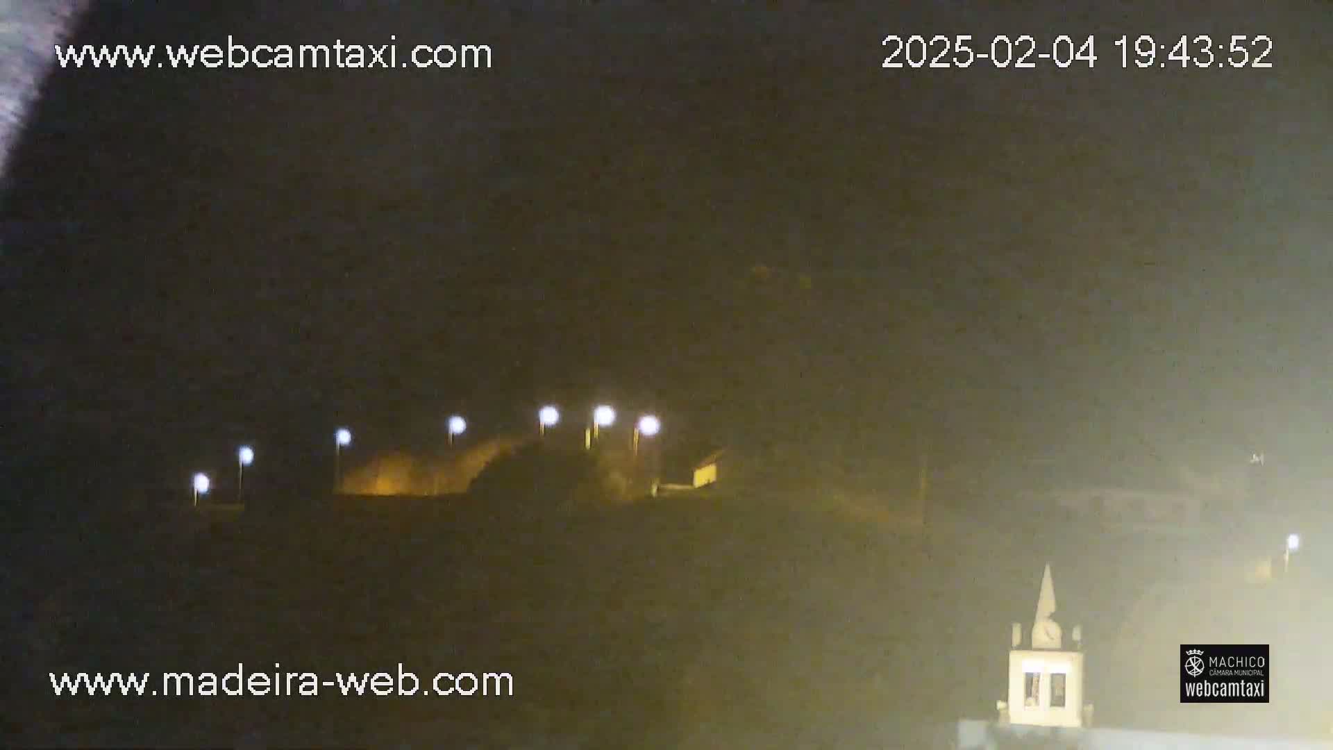 A nighttime, dimly lit outdoor scene shows a hilltop with several lights, and a small building with a clock tower in the lower right corner.