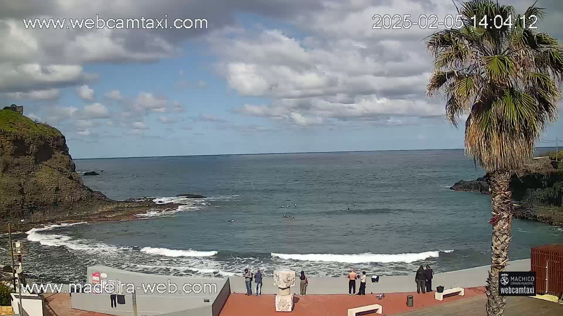 A partly cloudy day at a rocky ocean cove shows several people standing near the water's edge, with small waves lapping the shore.