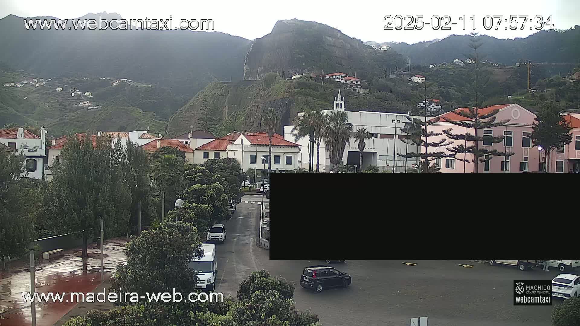 A wet, overcast day reveals a town nestled in a valley between hills, with several cars parked in a lot and some driving on the street.
