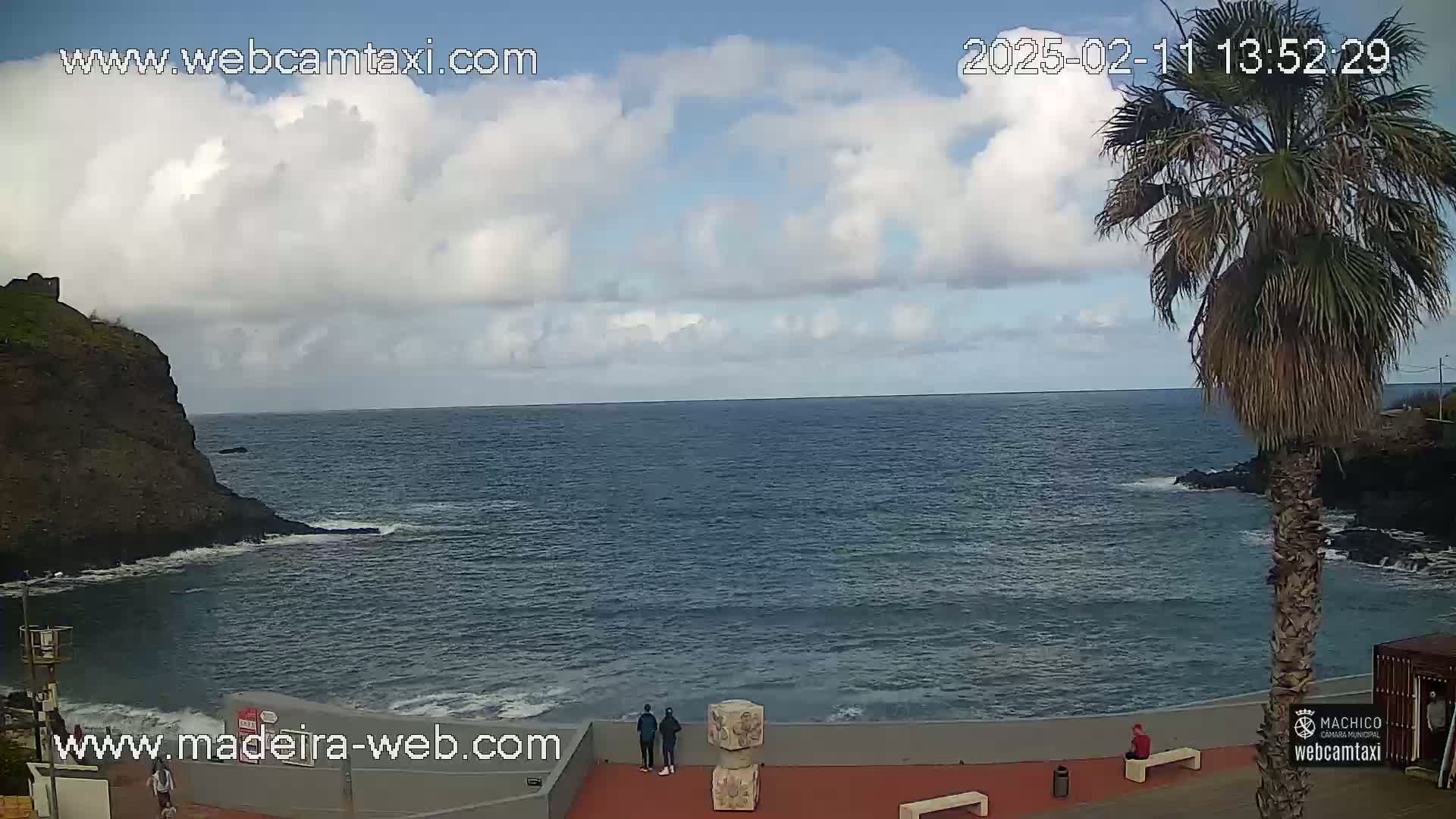 A partly cloudy day shows a rocky coastline with dark blue ocean waves, a palm tree, and a few people near a stone structure on a paved area.
