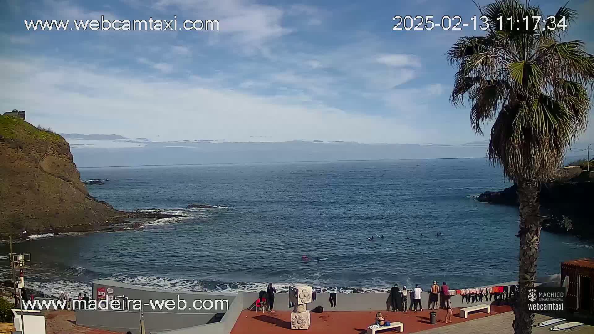 A mostly sunny day shows a rocky cove with calm ocean water, several people gathered on a beach, and a palm tree in the foreground.