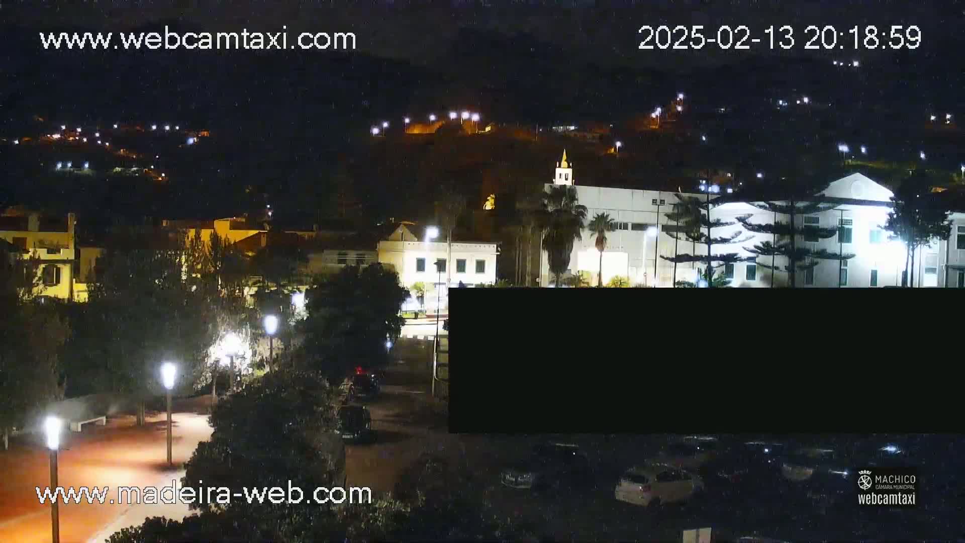 A nighttime view of a town with illuminated buildings and streets, cars parked in a lot, and palm trees.