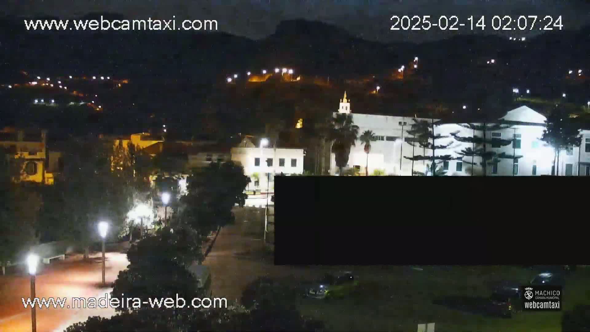 A nighttime view of a town nestled against a hill, illuminated by streetlights and building lights under a dark sky.