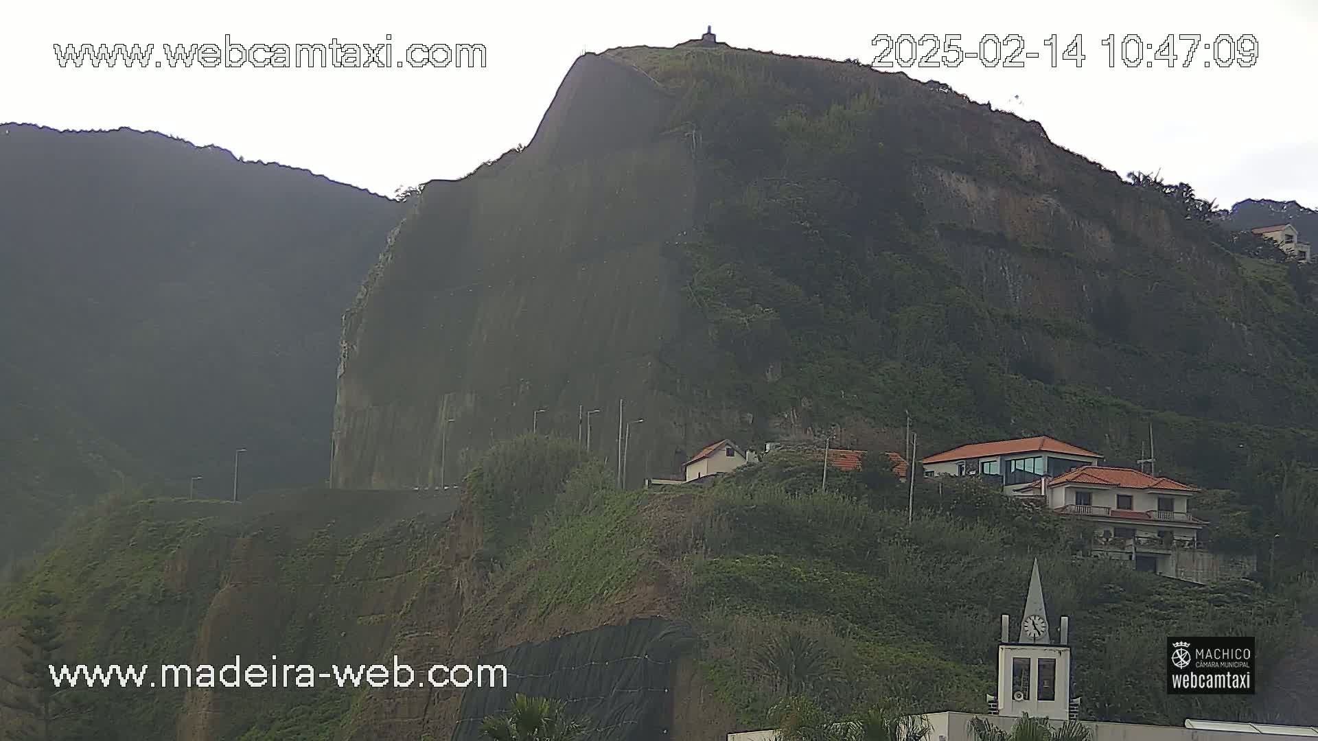 A large, dark-green cliff, partially shrouded in mist, overlooks several houses and a small church, nestled amongst lush vegetation on a hillside.