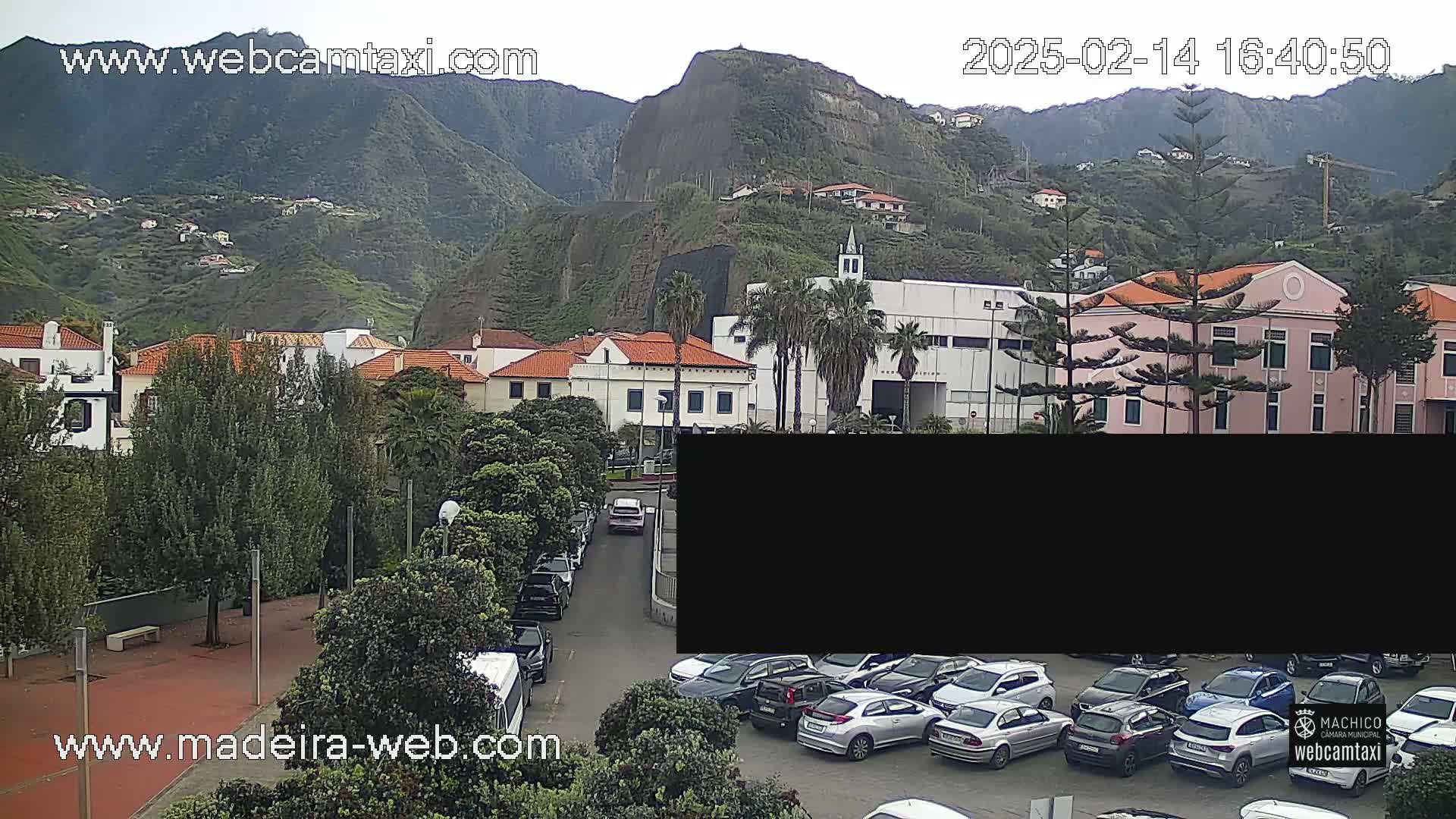 A street-level view of a town nestled against a mountainside on an overcast day, showing buildings, parked cars, and lush green vegetation.
