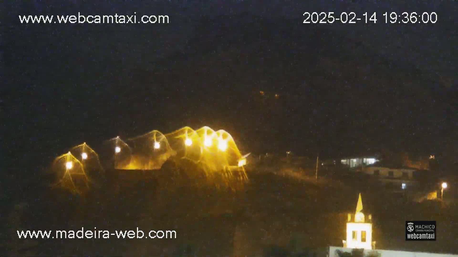 At night, illuminated structures on a hill are visible, with a small building and faint lights in the distance under a clear, dark sky.