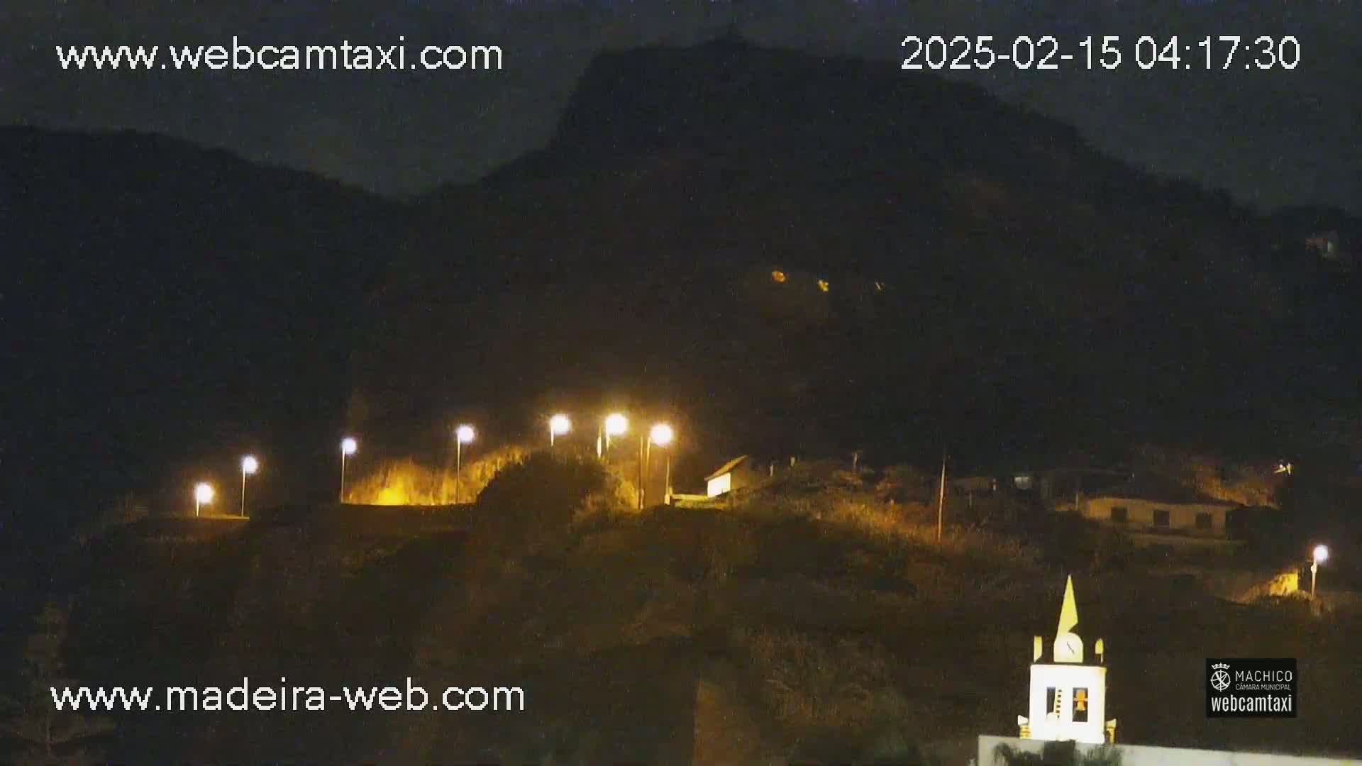 A nighttime view of a hilltop village illuminated by streetlights, nestled against a dark mountain under a clear night sky.