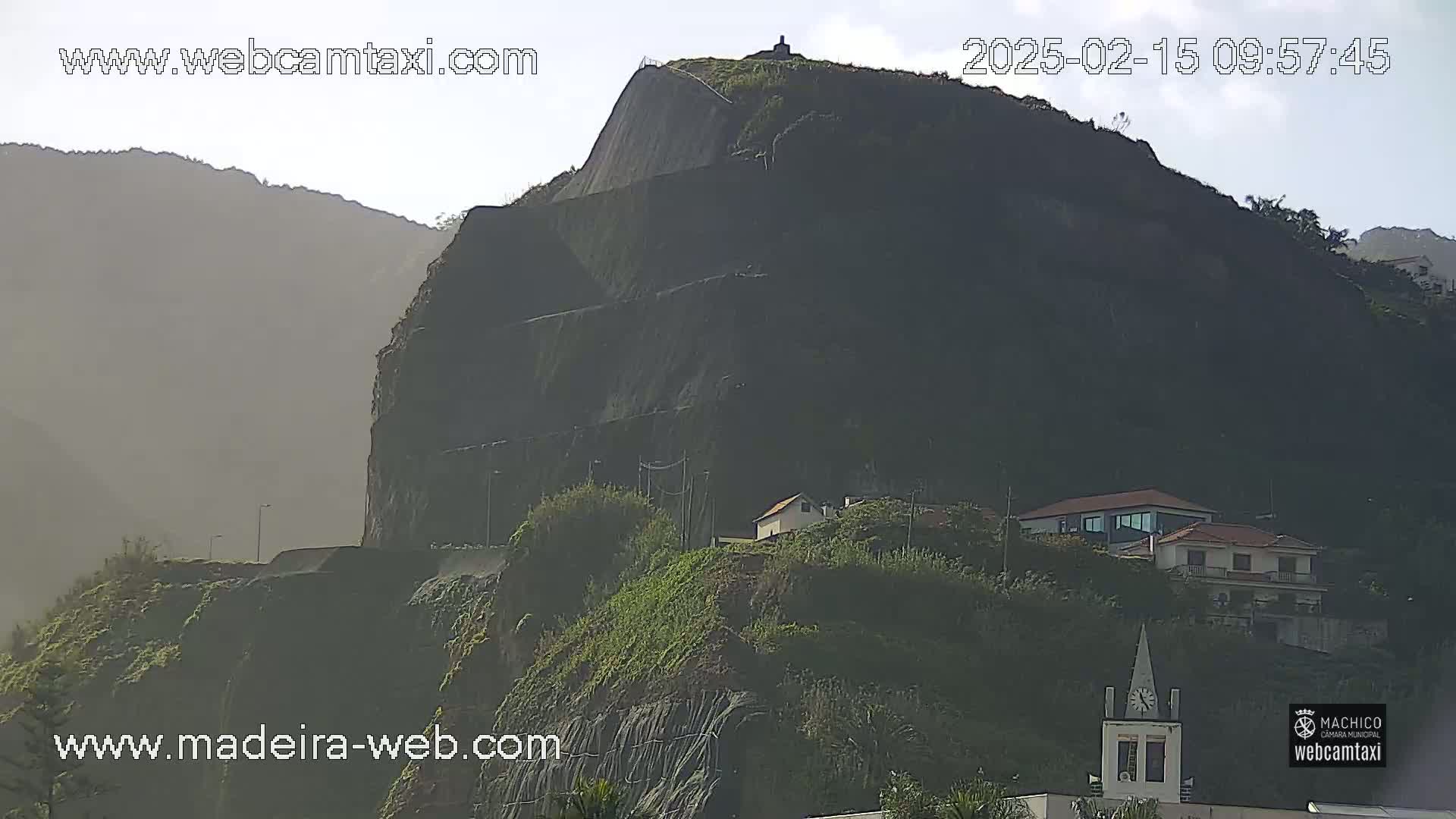 A large, dark, terraced hillside with a few buildings nestled into the greenery, under a mostly clear sky.