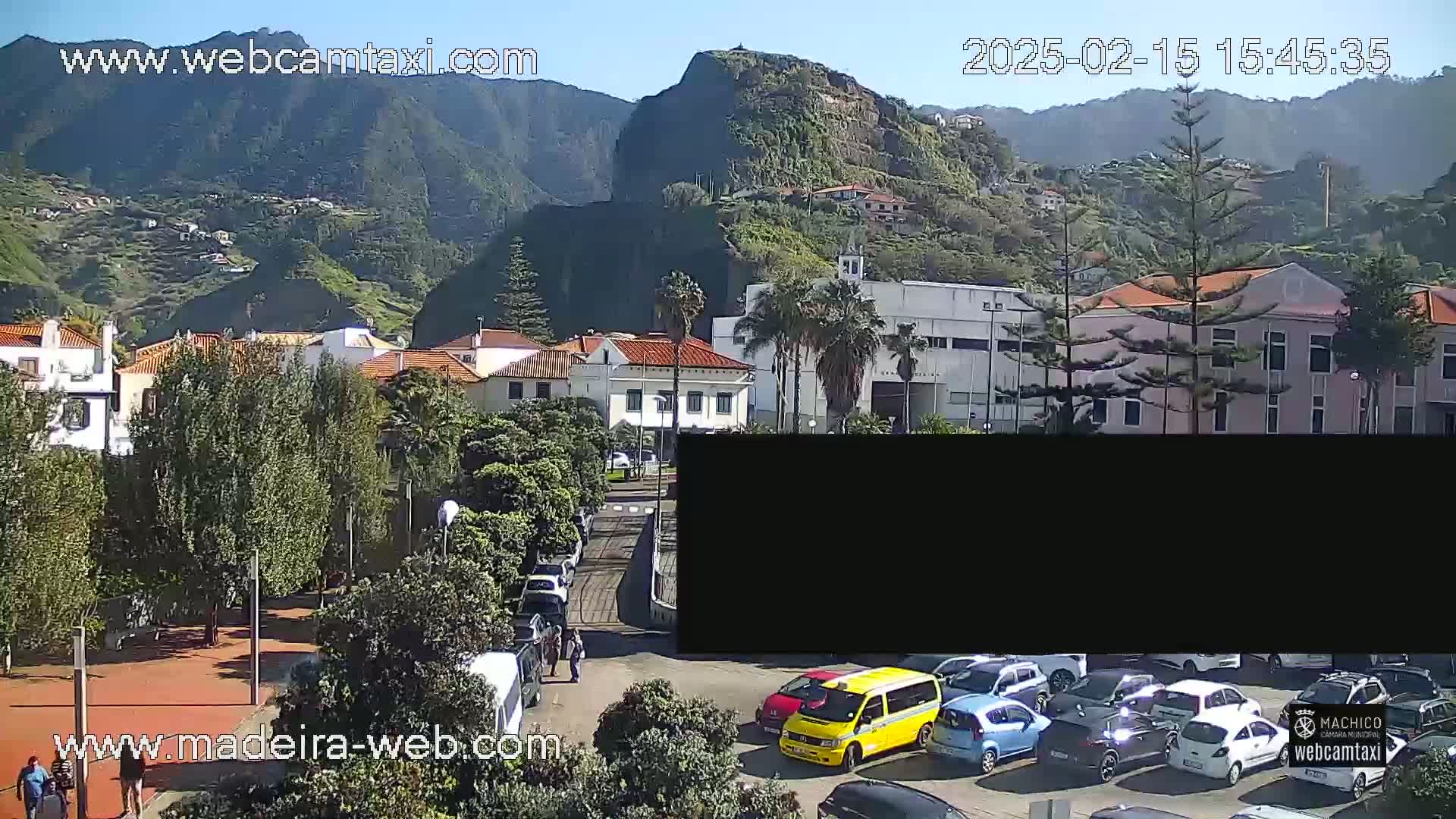 A sunny view of a town nestled against a mountainous backdrop, with cars parked in a lot in the foreground.