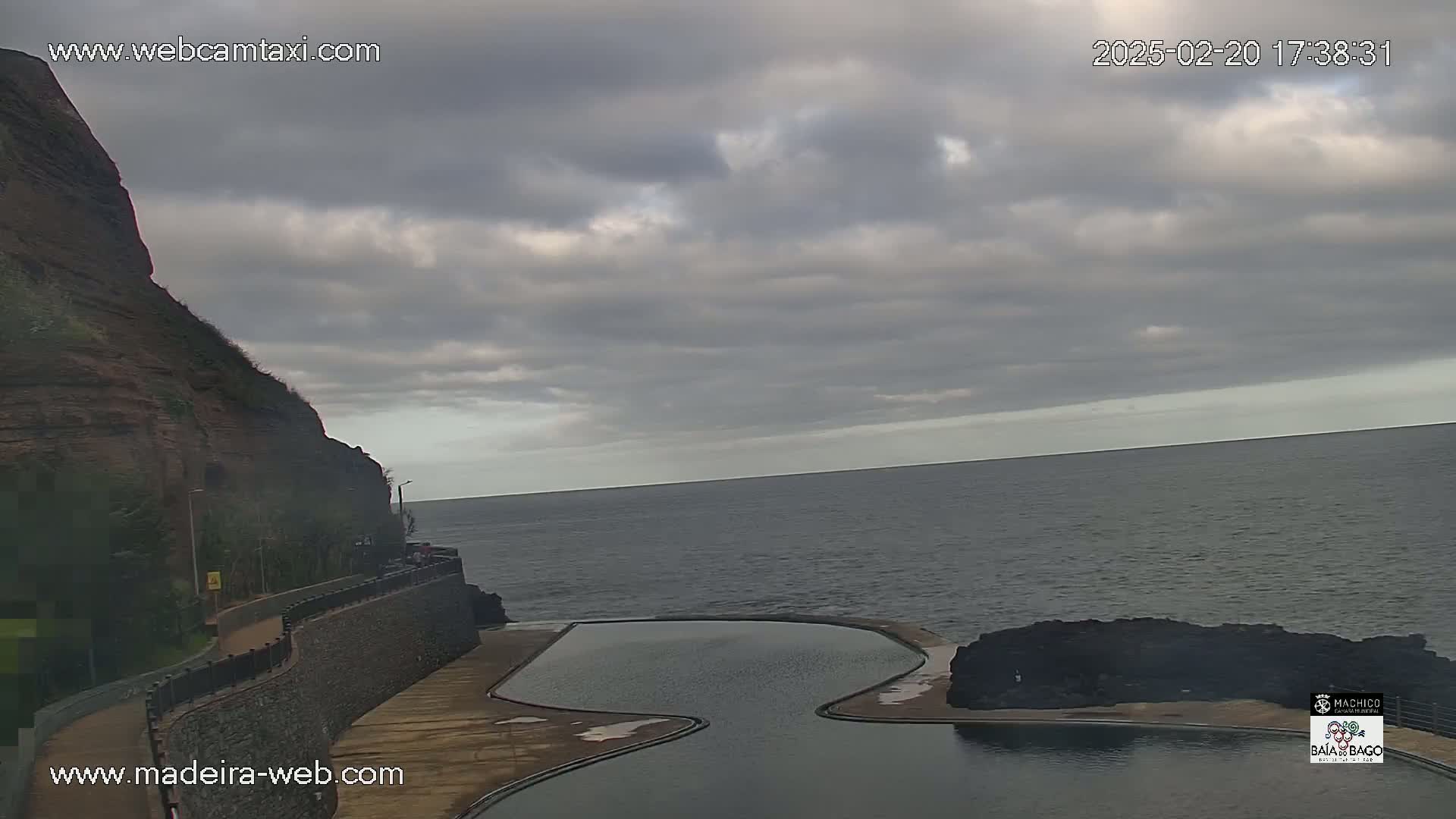 An oceanside, kidney-shaped pool sits beside a rocky cliff under a mostly cloudy sky.
