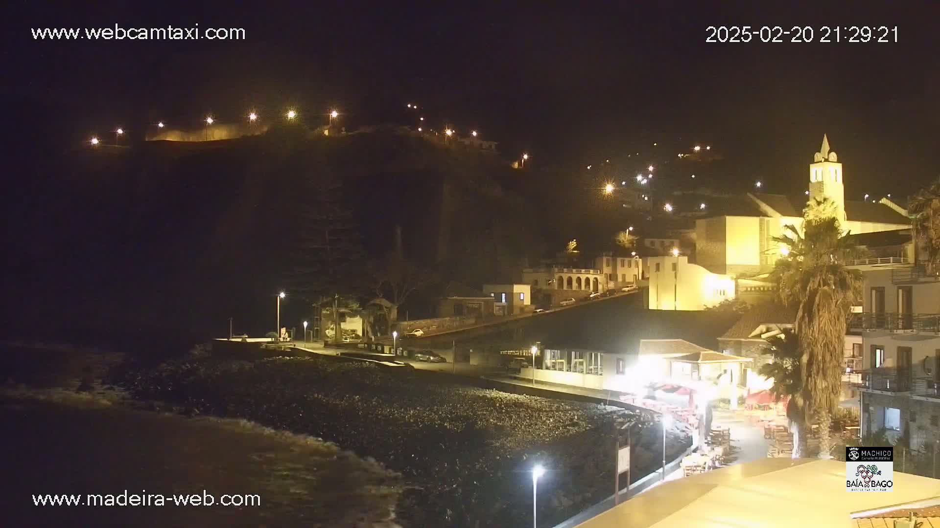A nighttime coastal town scene with a church, buildings, and palm trees illuminated by streetlights, with gentle waves lapping at a dark beach.