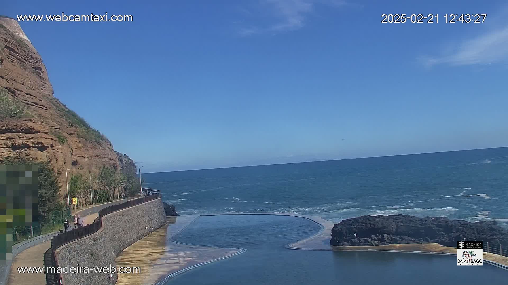 A rocky cliff overlooks a calm ocean with a wave-cut pool next to dark volcanic rock formations under a clear blue sky.
