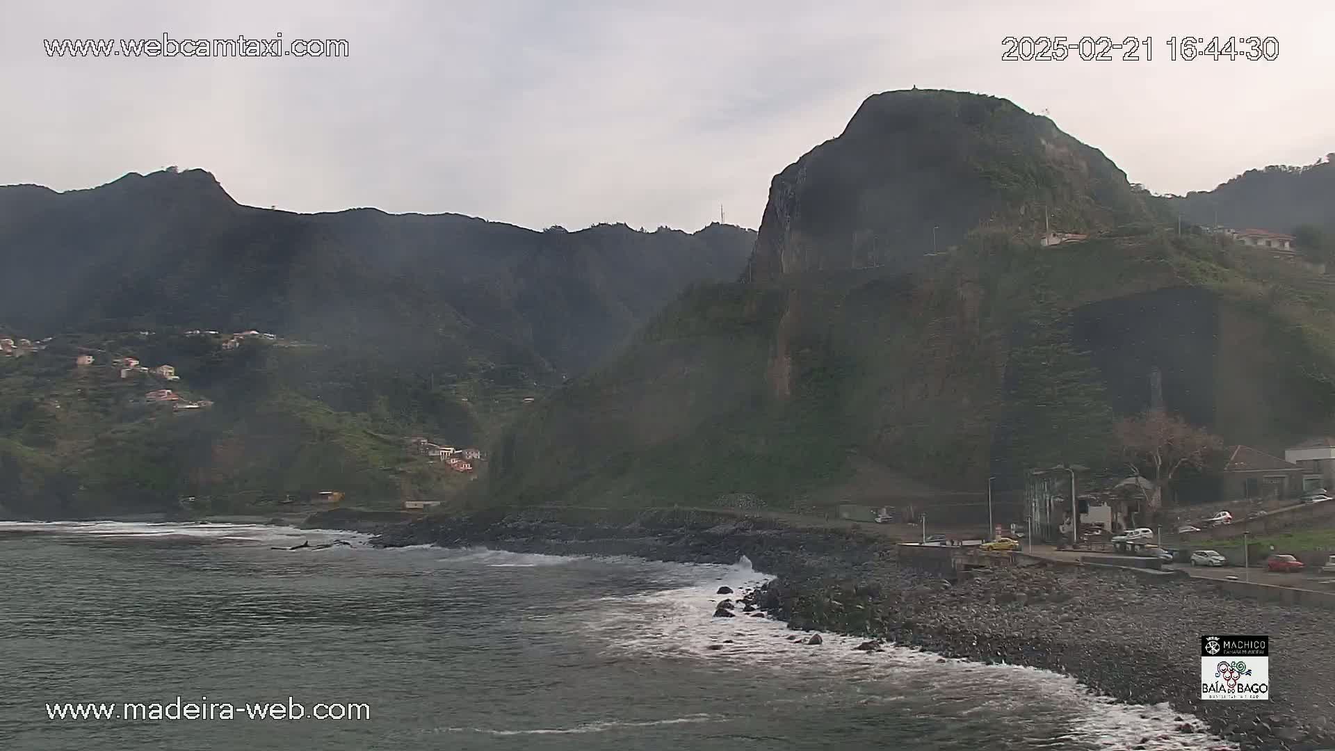 A rocky coastline with a village nestled in dark green mountains under an overcast sky, shows gentle waves lapping the shore.