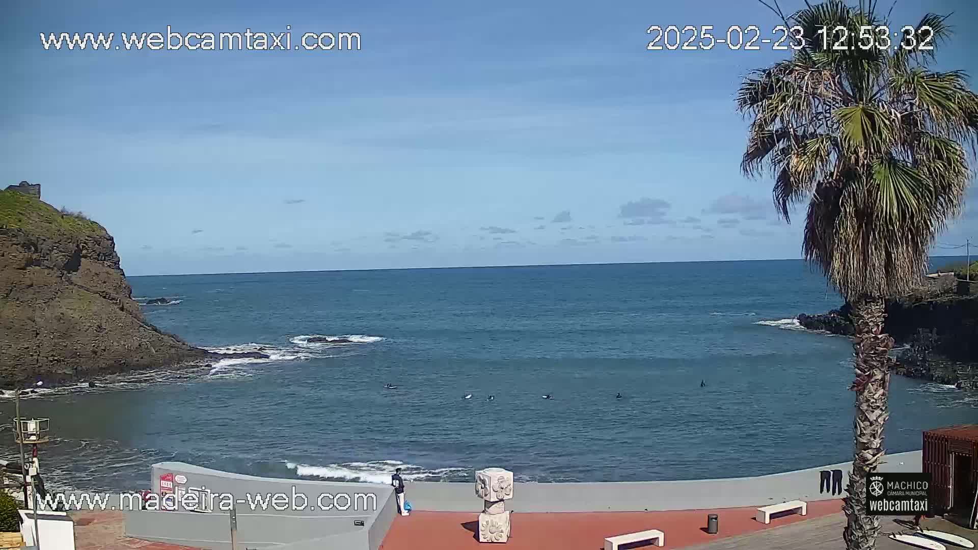 A calm ocean cove with several people in the water, viewed from a seaside promenade under a mostly sunny sky.