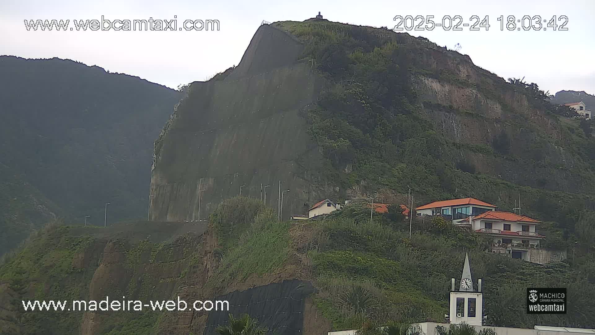 A large, dark-colored cliff face, partially covered in vegetation, overlooks a few houses and a clock tower under an overcast sky.