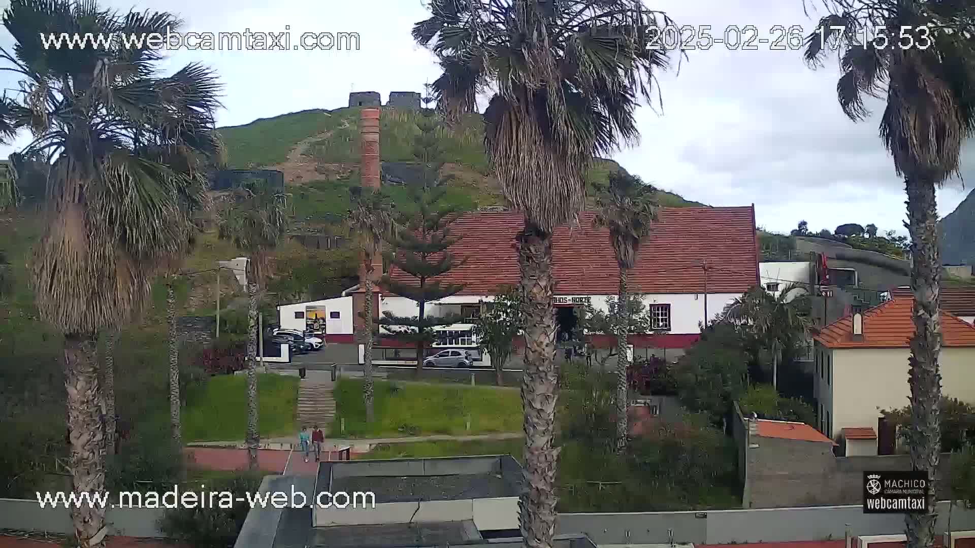 A partly cloudy day reveals a seaside town nestled below a hill with a stone structure, palm trees swaying in the breeze, and several buildings with red-tiled roofs.