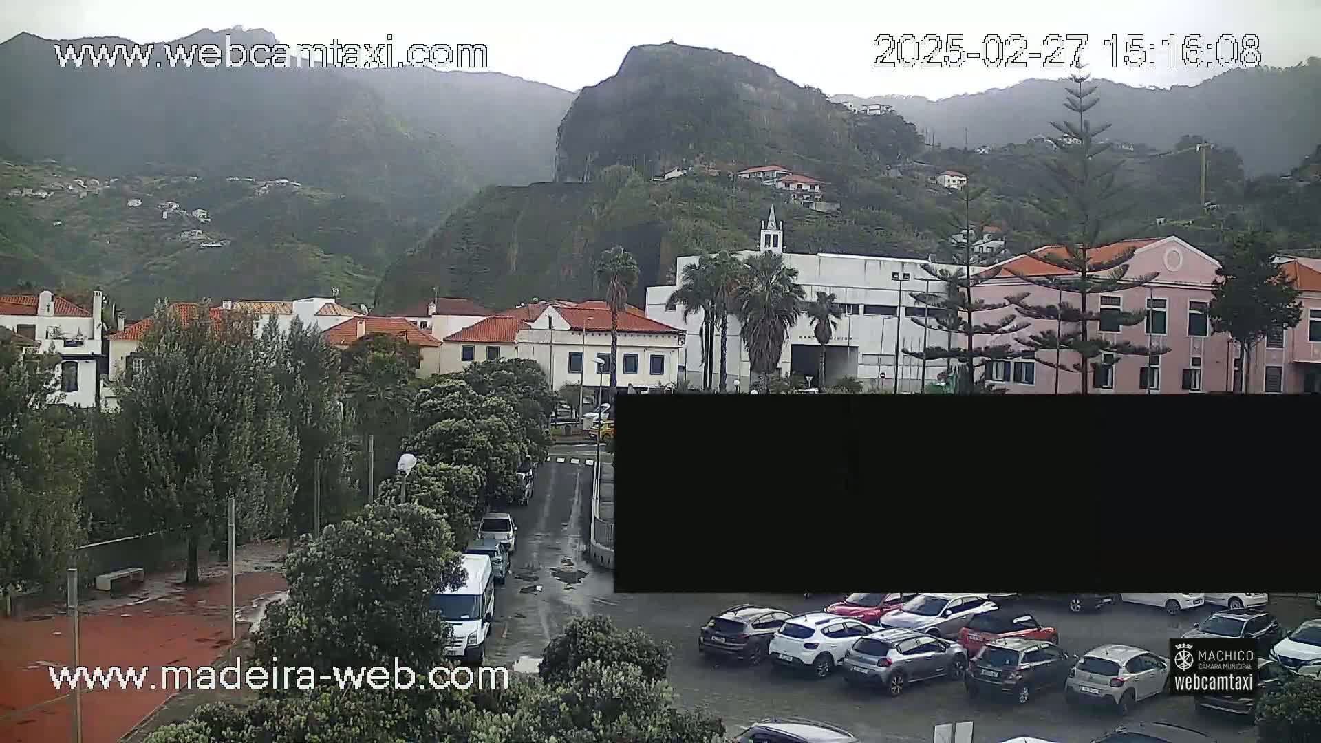 A rain-swept view of a town nestled against a verdant mountainside, with cars parked in a lot in the foreground.