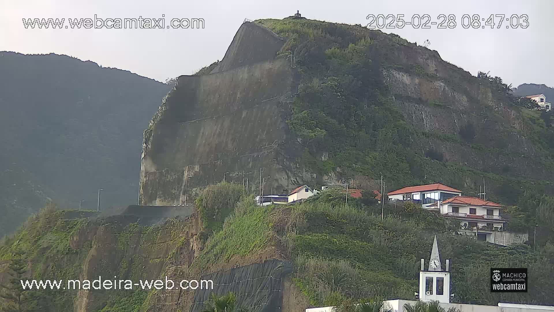 A large, steep, green hill with a road and houses at its base is shown under overcast skies.