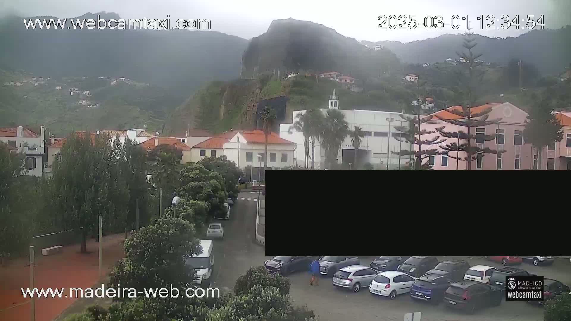 A misty, overcast day reveals a parking lot filled with cars in front of several buildings nestled against a hillside, with a prominent rock formation visible in the background.