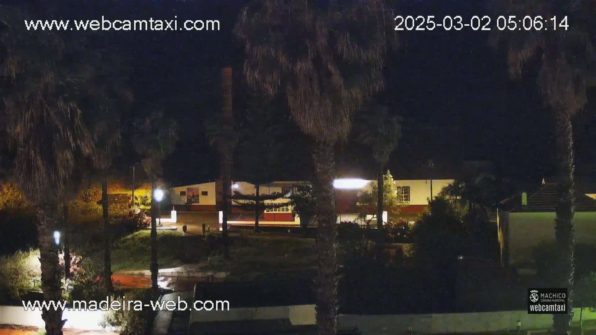 At night, palm trees frame a view of a low-lit building complex with a paved area in front.
