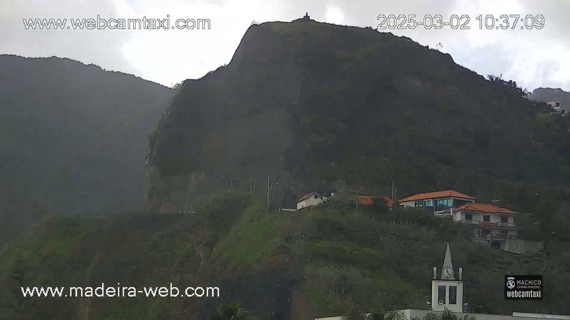 A large, dark-green mountain with a few buildings on its lower slopes is shown under an overcast sky.
