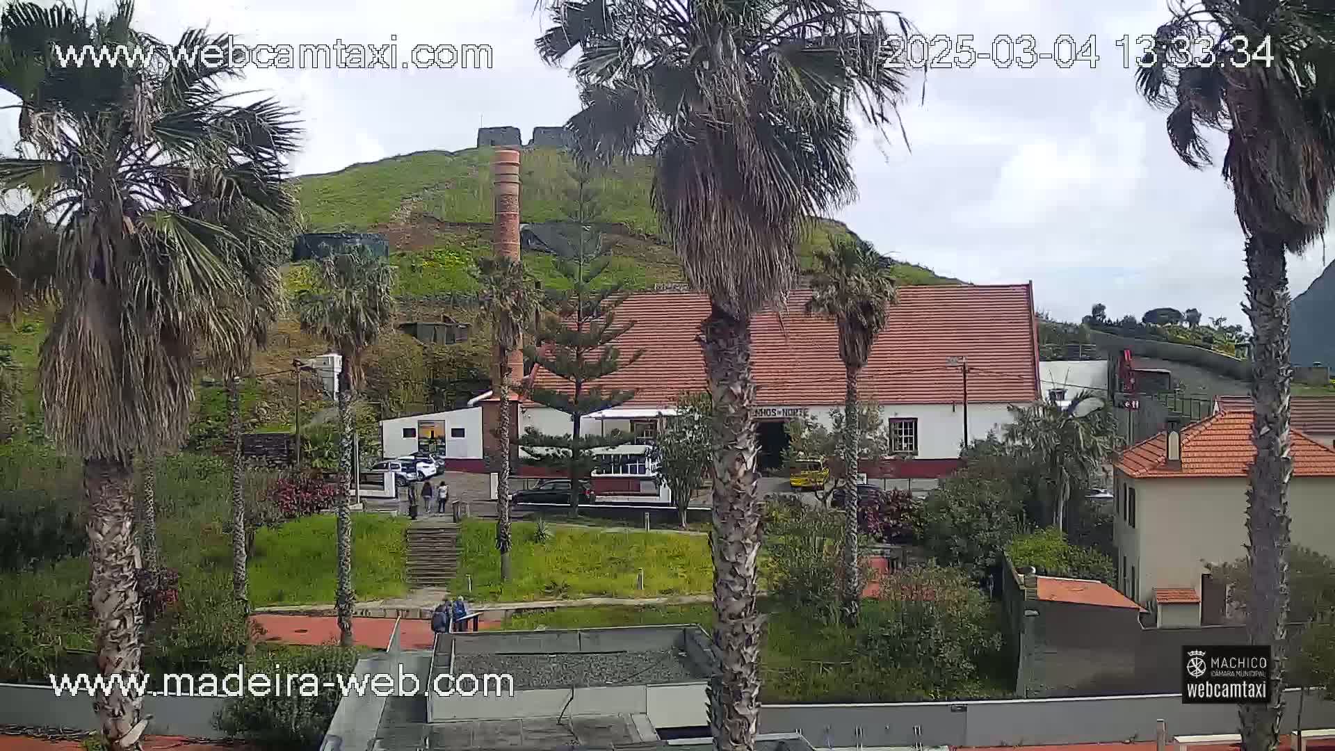 A partly cloudy day reveals a hillside town with palm trees, a white building with a red roof, and a brick chimney.