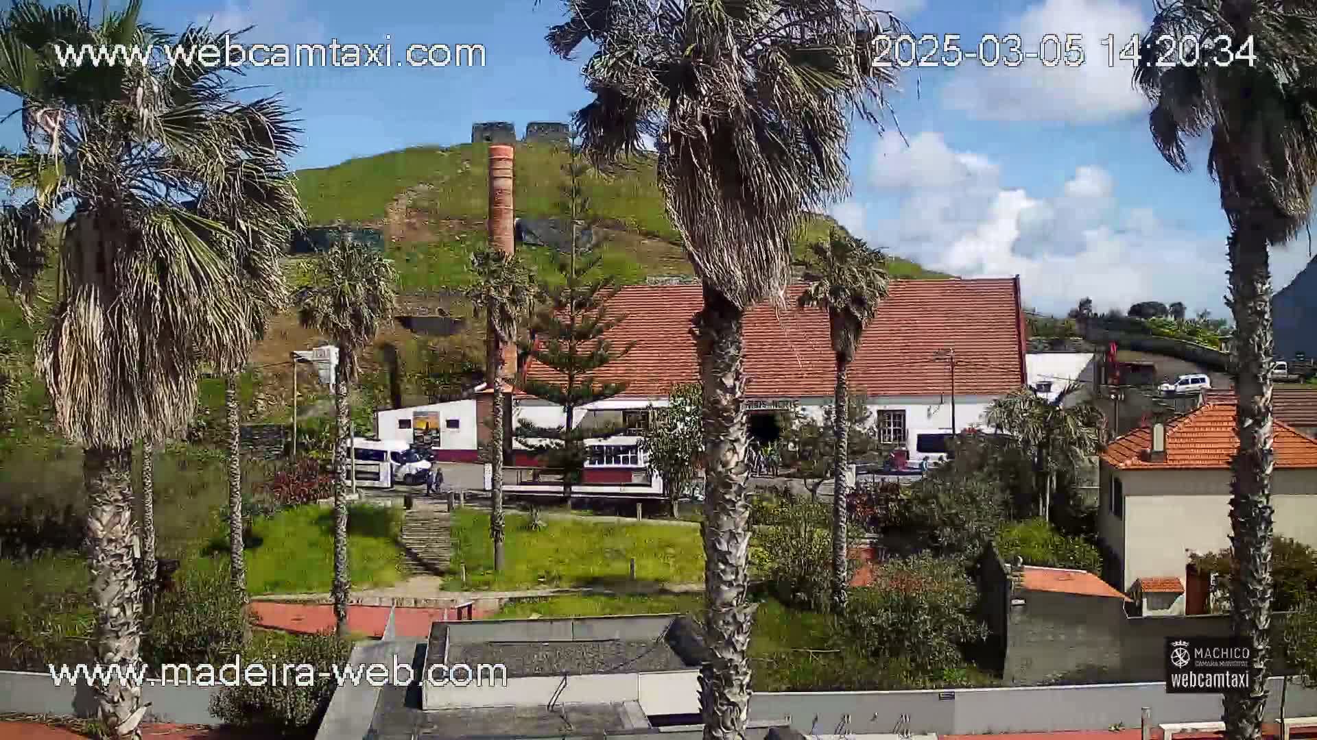 A partly sunny day reveals a red-roofed building near palm trees and a hill with old brick structures, all overlooking a street with parked cars.