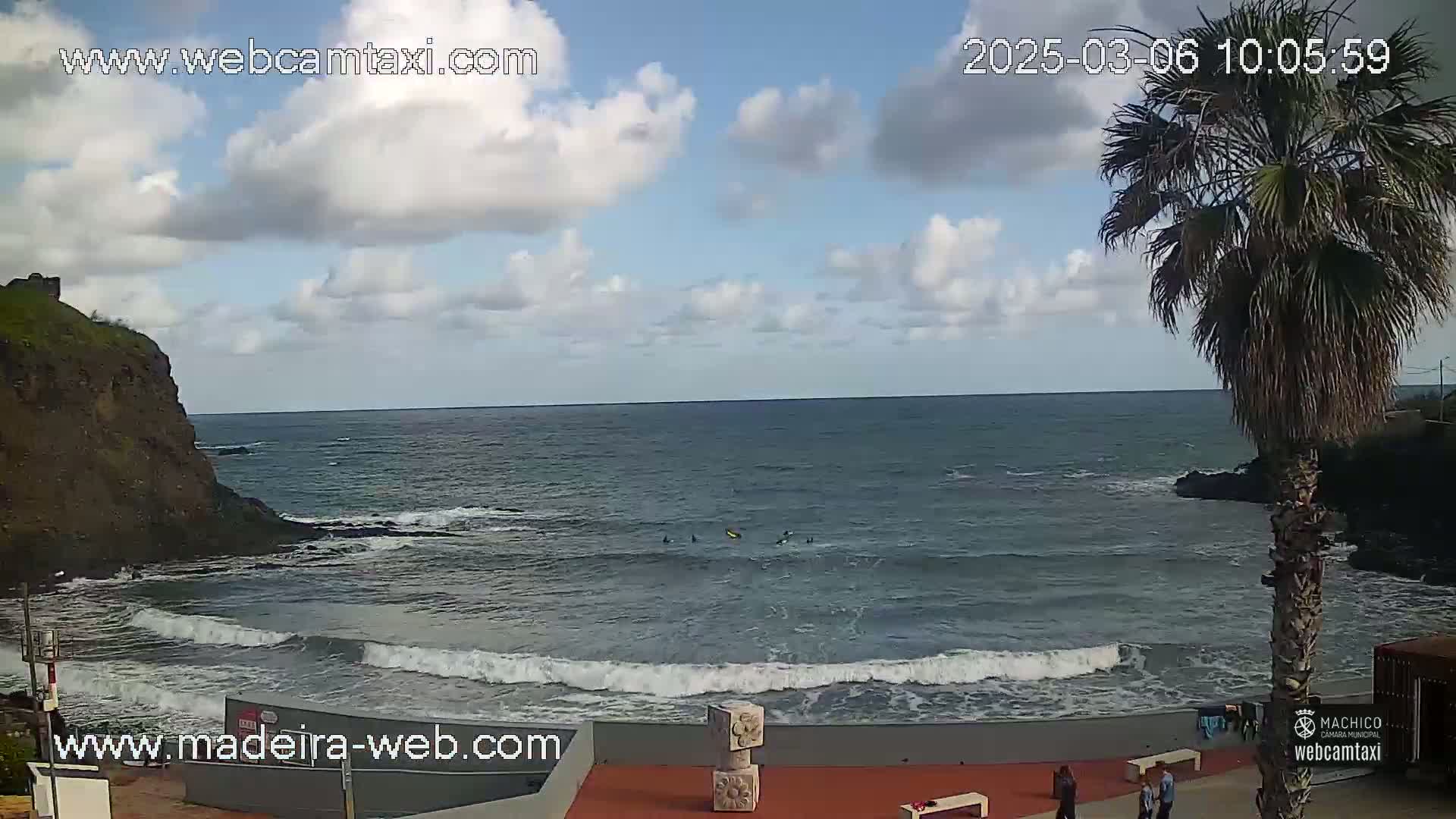 A partly cloudy day shows a rocky coastline with a small beach where several people are in the water, a palm tree stands near a red paved area.