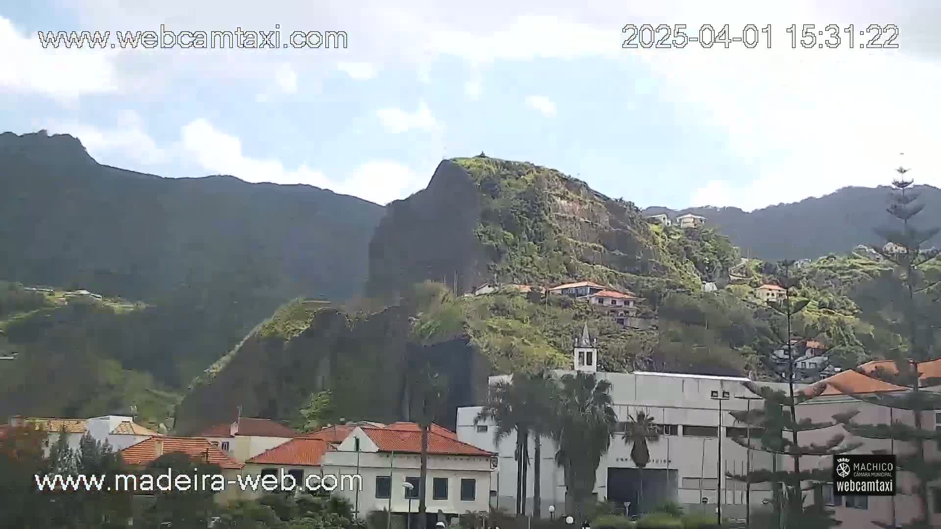 A partly cloudy day reveals a hillside village nestled against steep, dark volcanic cliffs, with palm trees and buildings visible in the foreground.