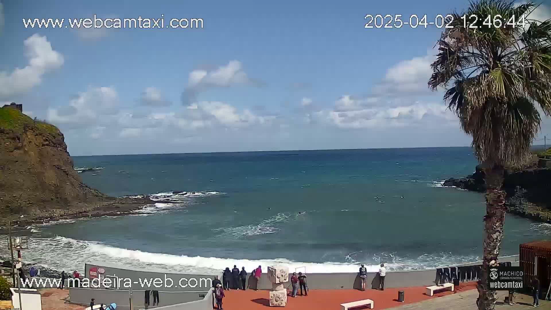 A rocky cove with a calm ocean under a partly cloudy sky, where people stand on a wall overlooking the water.