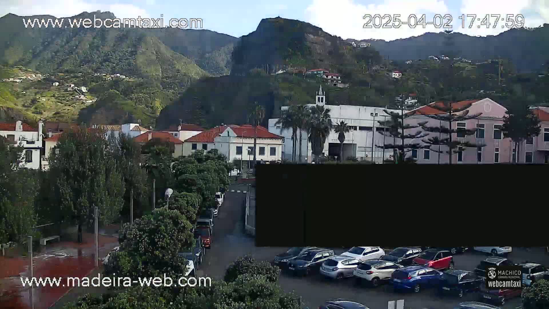 A town nestled against a verdant mountainside under partly cloudy skies, with cars parked in a lot in the foreground.