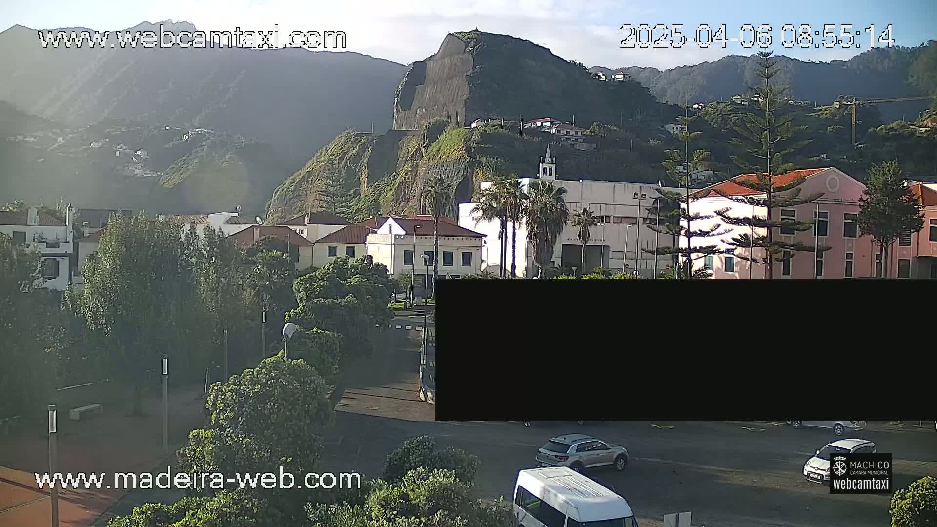 A sunny outdoor view of a town nestled against a steep, rocky mountainside, with parked cars visible in a parking lot below.