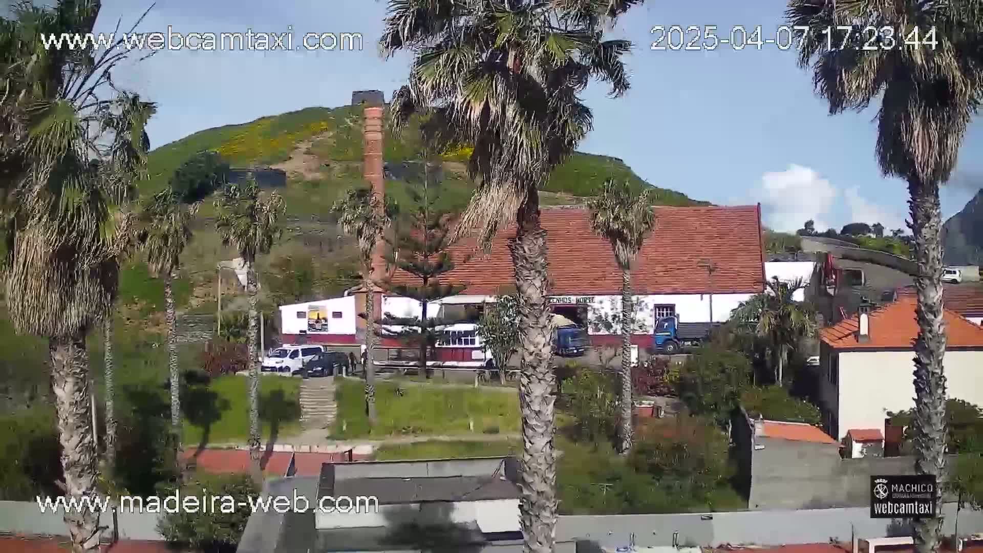 A sunny day reveals a hillside village with palm trees, low buildings, and a larger structure with a smokestack, near vehicles parked in front.
