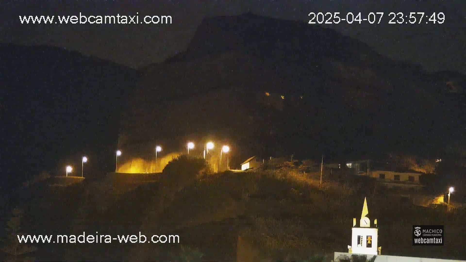 A nighttime view of a hilly landscape with illuminated buildings and a church steeple, under a clear, dark sky.