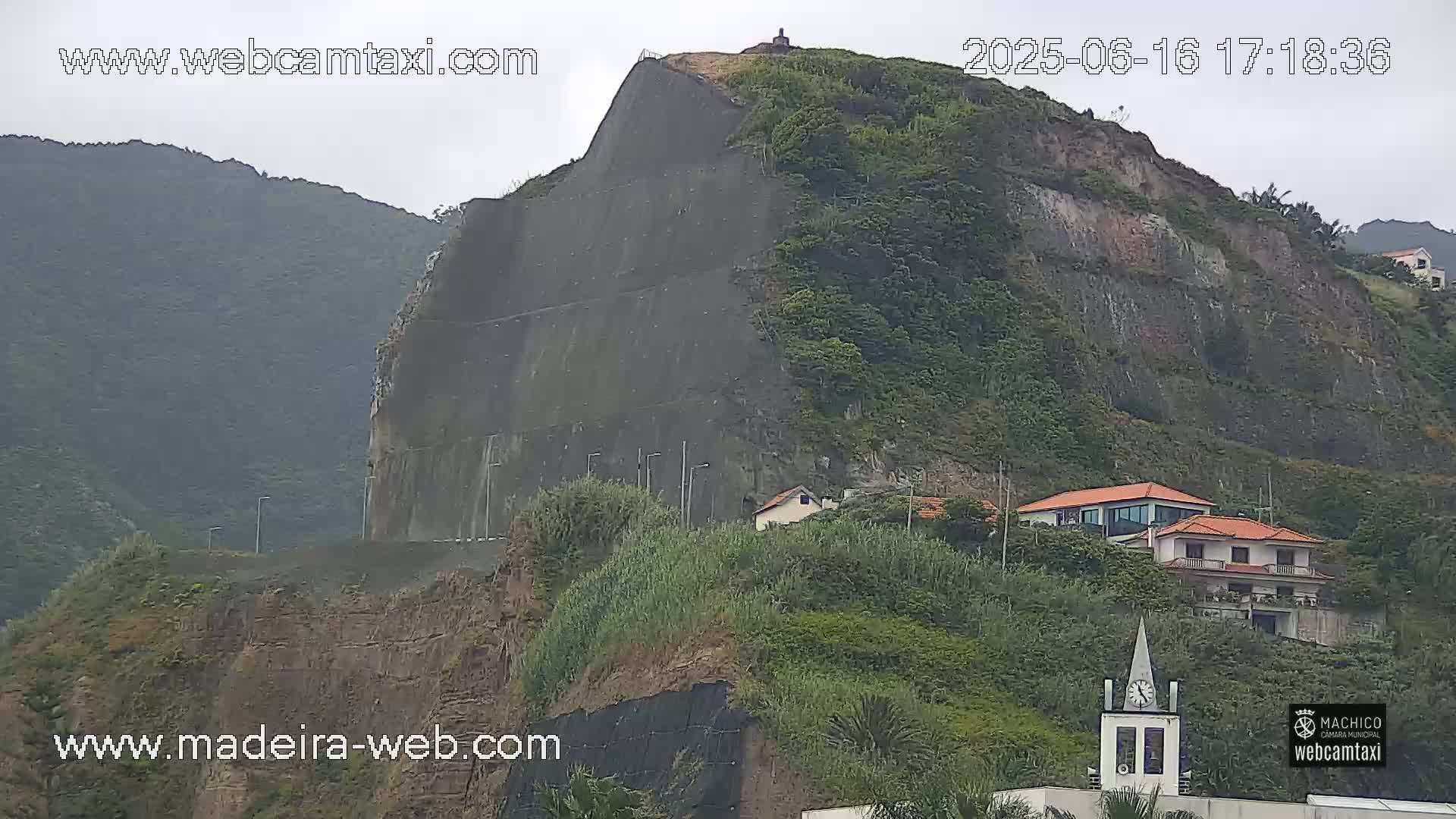 A large, dark gray retaining wall is built into a steep, green hillside under an overcast sky; a few houses are visible at the base of the hill.