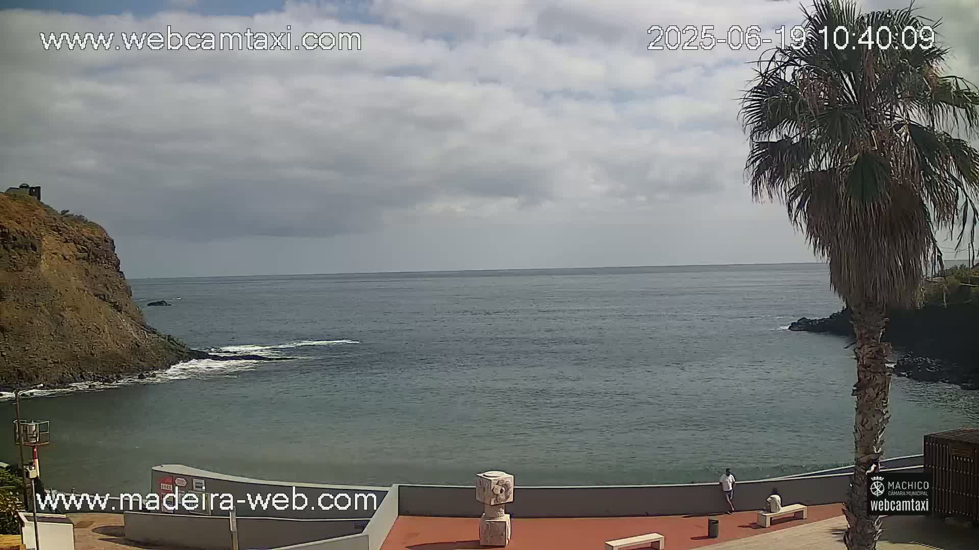 A partly cloudy day shows a calm ocean cove next to a rocky cliff, with a palm tree and a few people visible on a nearby promenade.