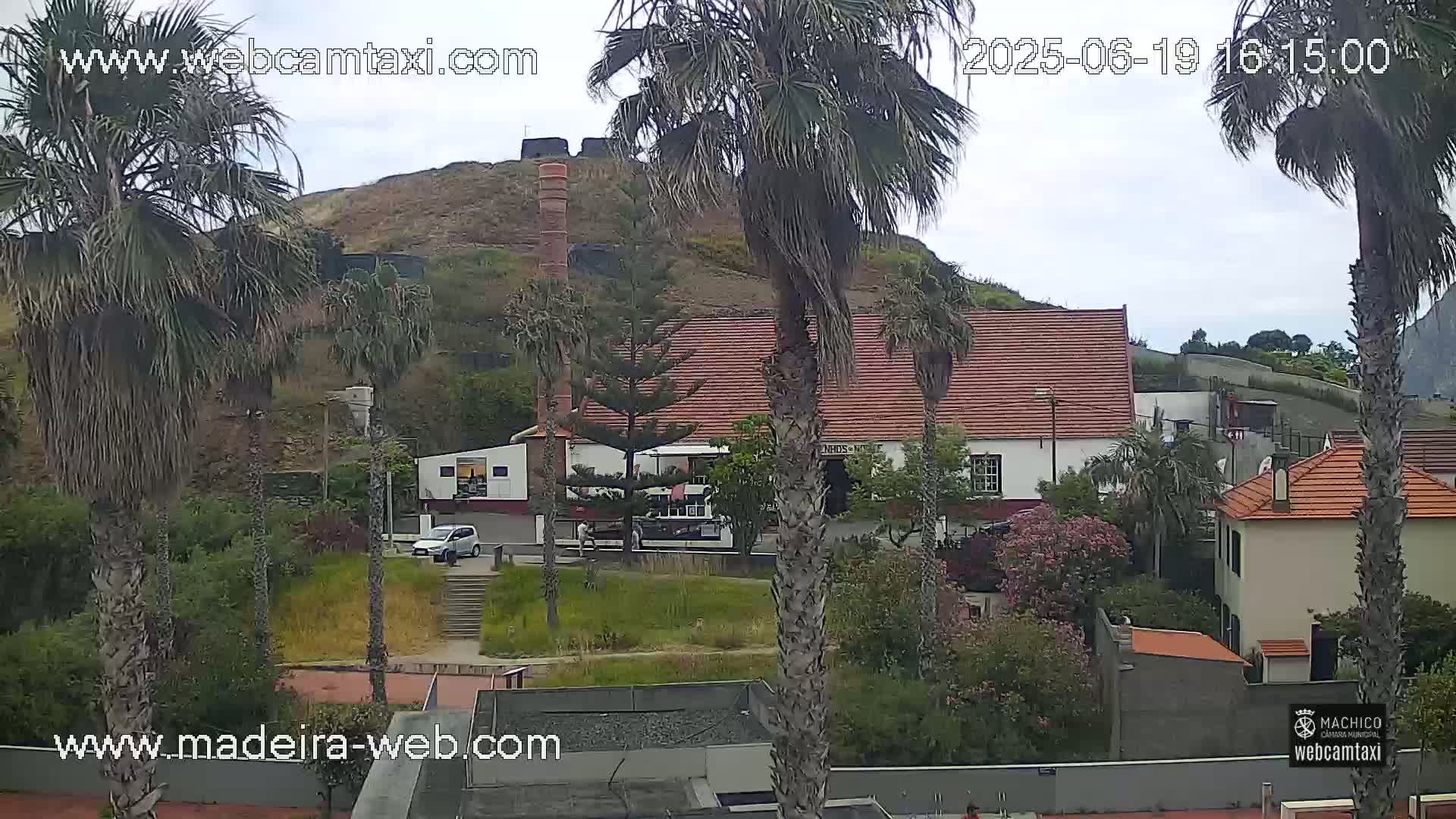 A slightly overcast day reveals a hill with a brick structure, buildings with red tile roofs, palm trees, and lush vegetation.