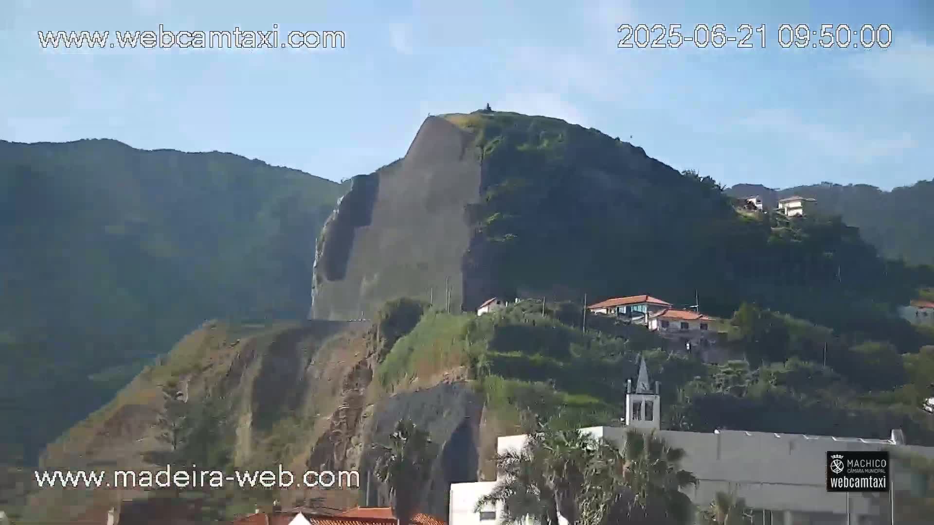 A rocky, green hill with houses and a church on its lower slopes is seen on a clear, sunny day.