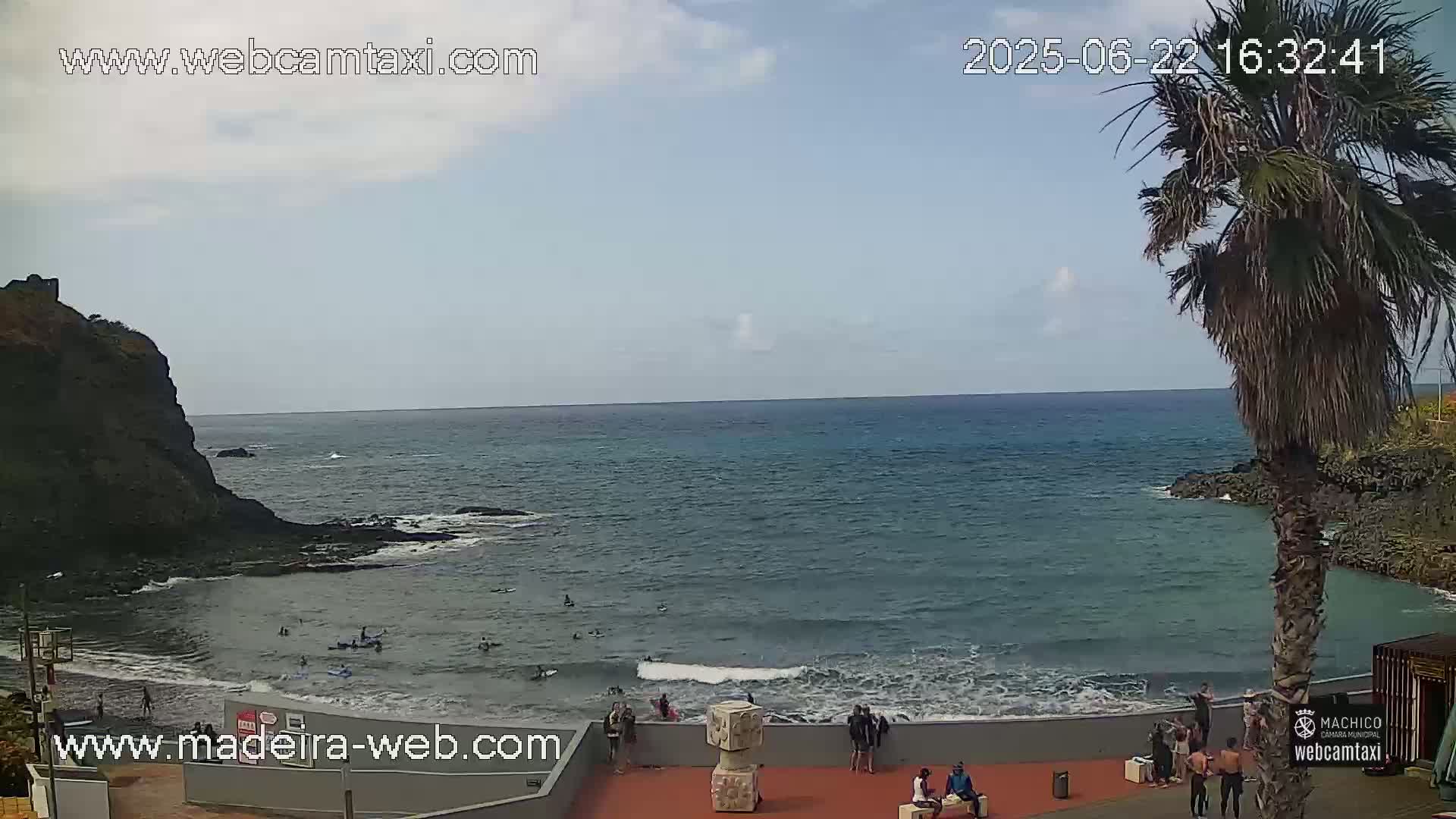 A partly cloudy day shows a calm ocean with people swimming and standing on the shore near a rocky cliff and palm trees.