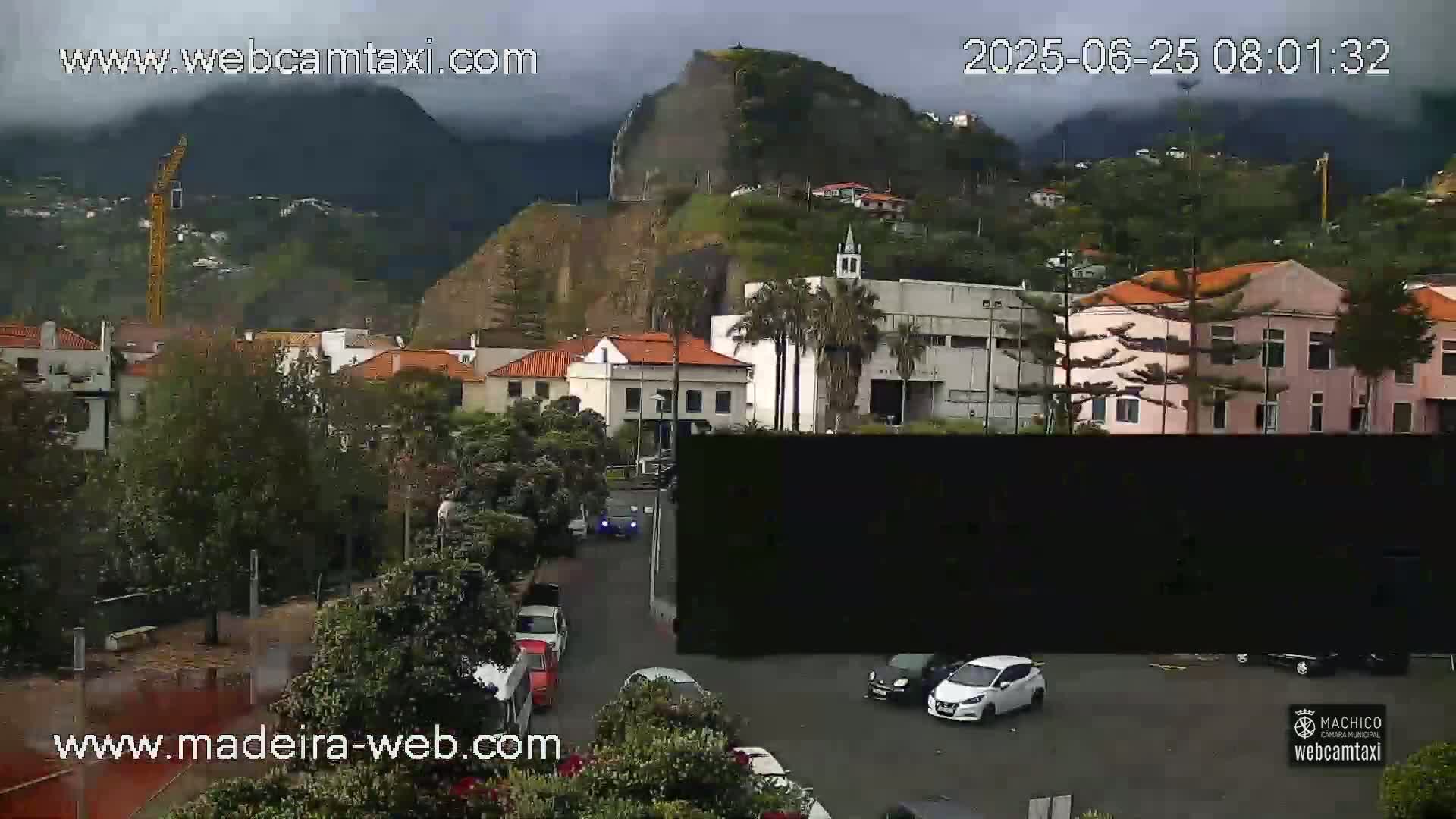 A cloudy day reveals a coastal town nestled below a steep, green hill, with several cars parked in a lot and a few more driving on the street.