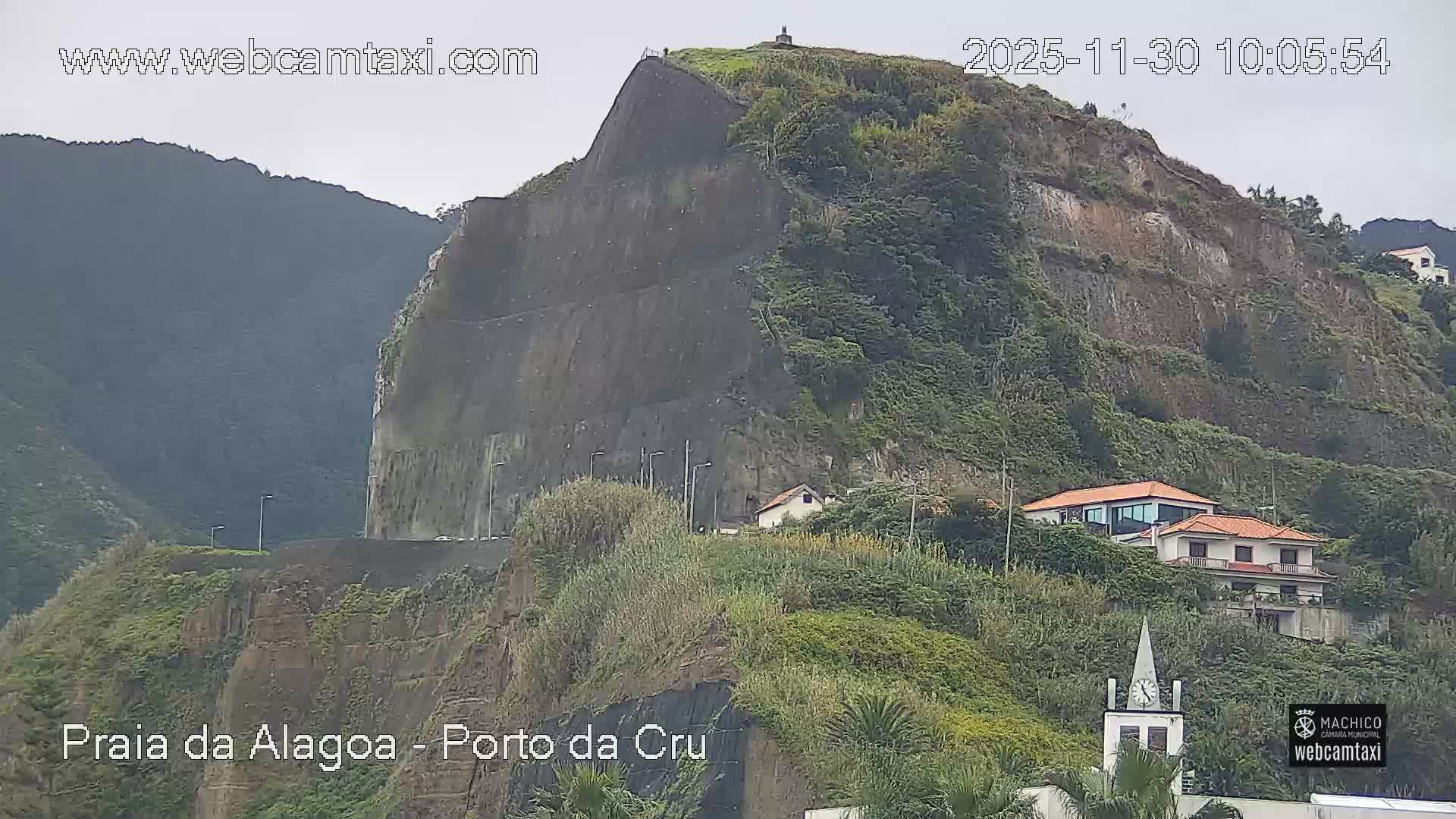 Under an overcast sky, a large, steep mountain with a mixture of lush green vegetation and terraced, exposed rock faces is dotted with a winding road, several houses, and a prominent church steeple, with distant hills visible on the left.
